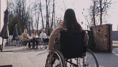 Vista-Trasera-De-Una-Mujer-Discapacitada-En-Silla-De-Ruedas-Que-Se-Encuentra-Con-Otros-Amigos-En-Una-Terraza-De-Bar