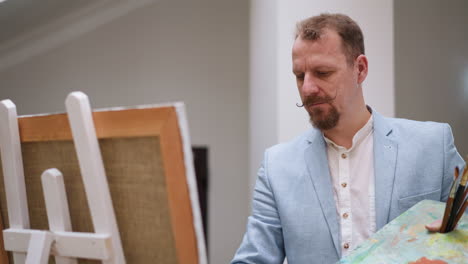 el creador de arte dibuja en lienzo en el estudio. artista concentrado con barba y bigote sostiene una paleta rectangular de pie junto al caballete. pintor trabajando en la tienda