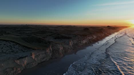 sea waves are washing the beach, high dunes have formed, a summer house is standing on the dunes, and the sun is setting on a winter evening