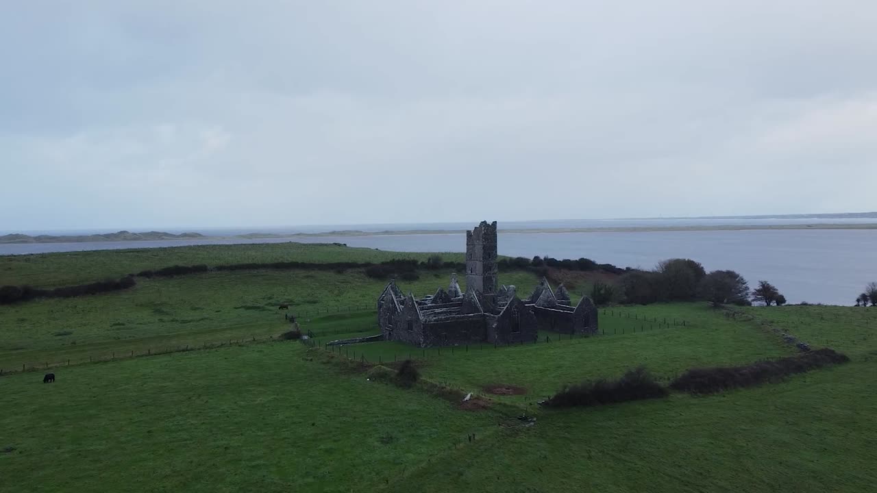 Luftfoto fra drone, der nærmer sig de historiske middelalderruiner af Moyne Abbey, smukt beliggende på et frodigt grønt irsk græsland ved kystfarvandene på en dyster, overskyet dag.