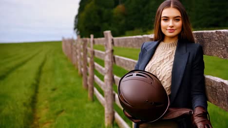 woman in stylish outfit leaning against a fence with a riding helmet