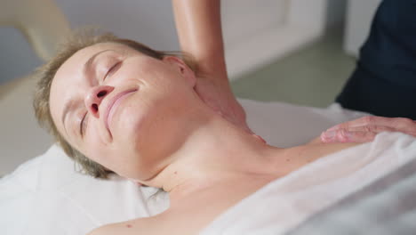 lady on massage bed smiles with eyes closed feeling deeply relaxed as skin care expert glides hands from neck to shoulders delivering gentle wellness treatment in serene spa environment
