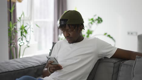 Hombre-Afroamericano-Negro-Usando-Un-Teléfono-Inteligente-Mientras-Descansa-En-Un-Sofá-En-La-Sala-De-Estar