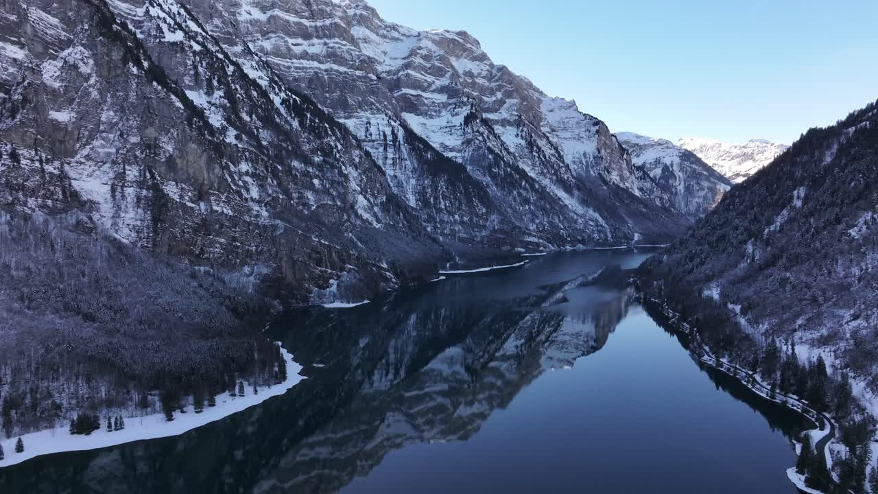 Drone beveger seg sakte bakover og avslører den speilblanke overflaten av Klöntalersee, mot et fjellrikt, snødekt bakteppe
