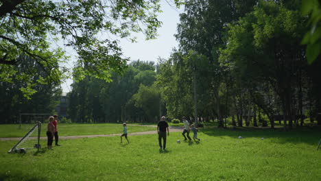 family enjoys an outdoor soccer game on a sunny day as the son in white dribbles past his brother in red and chases after the ball, which crosses the goal post