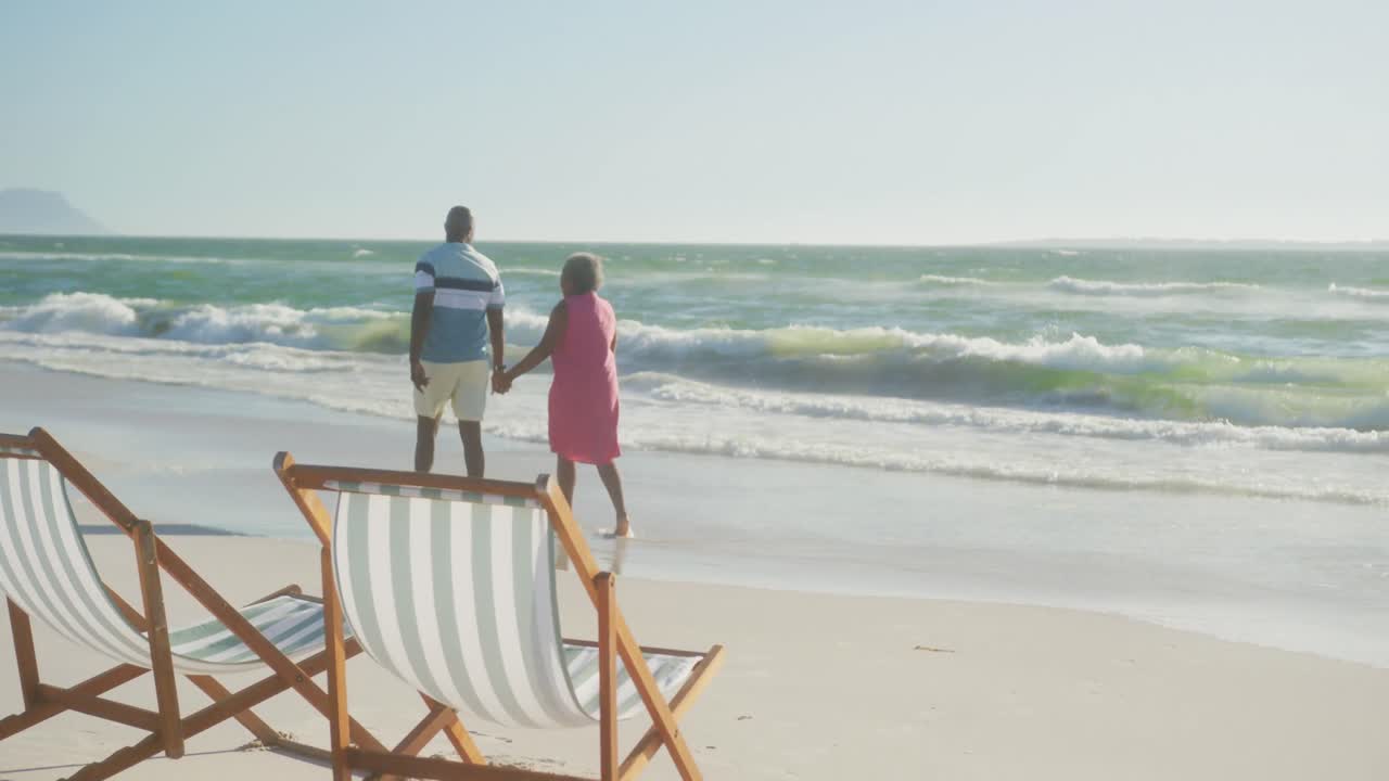 una feliz pareja de ancianos afroamericanos caminando y tomados de la mano en la playa, copia el espacio, en cámara lenta.