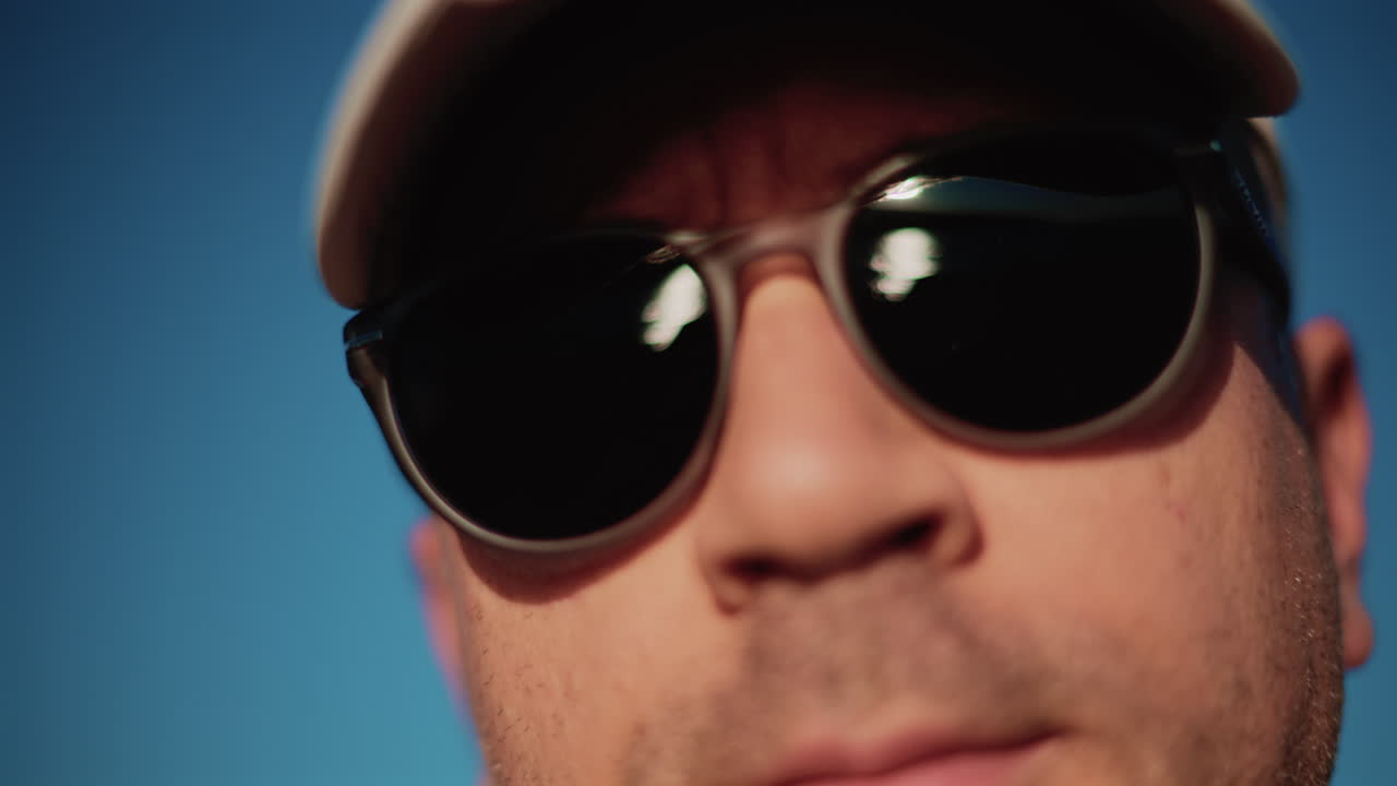 Extreme close up of a man wearing dark sunglasses and a cap, framed tightly against a clear blue sky in strong daylight