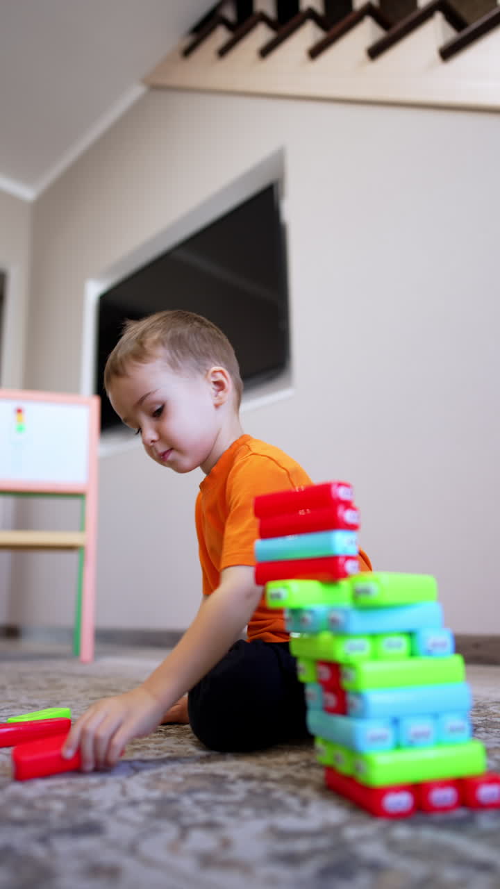 Small baby boy in orange t-shirt sits on the floor building a tower of blocks. Cute kid plays at home. Black cat at backdrop. Vertical video.