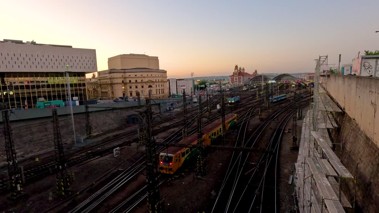 A yellow commuter train travels along multiple railway tracks toward a city station at sunset, with modern buildings and warm, ambient lighting