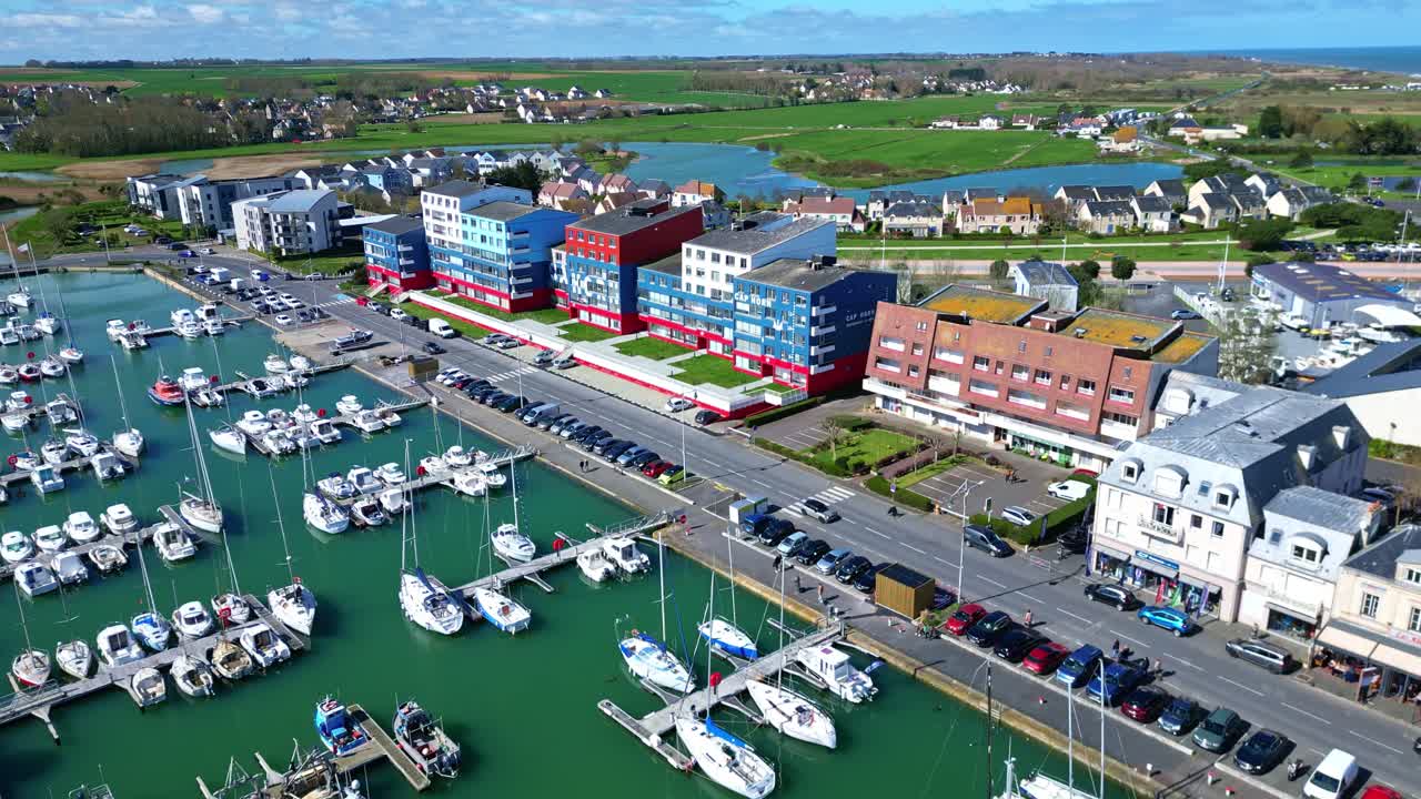 Forward drone shot of Courseulles-sur-Mer fishing port with seagulls, fields, traffic and colorful buildings. France