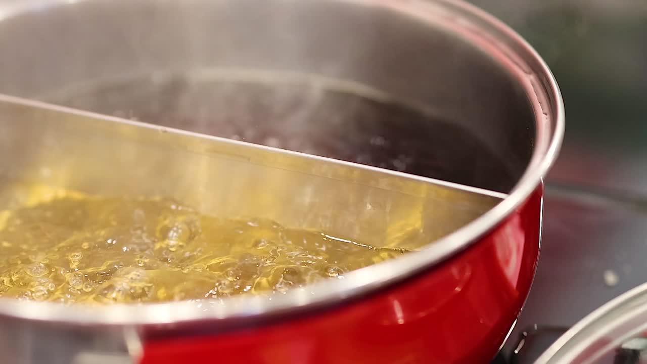 Close-up of a bubbling broth in a red hotpot, with steam rising and hands preparing ingredients.