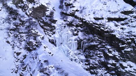 Aerial-close-view-of-a-frozen-waterfall-on-a-rocky-snow-covered-mountain-on-a-winter-day