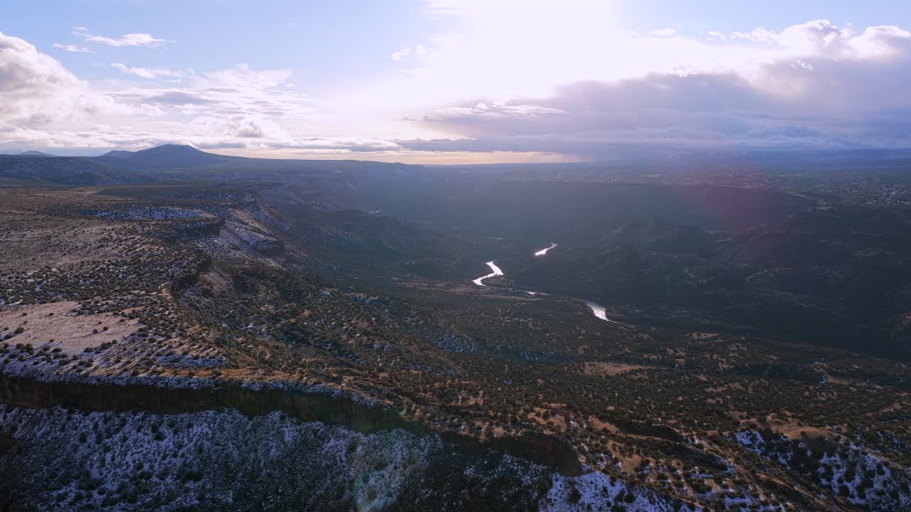 Una vista aérea cinematográfica capta un río sinuoso que brilla a la luz del sol bajo el borde de una meseta escarpada. La ligera nieve y las espectaculares nubes resaltan la vasta escala geológica de la naturaleza salvaje del Suroeste.