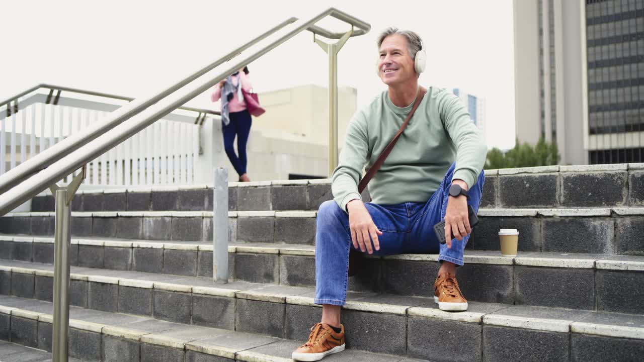 Arriving at plaza steps mature man sitting pulling headphones and tapping phone app while smiling