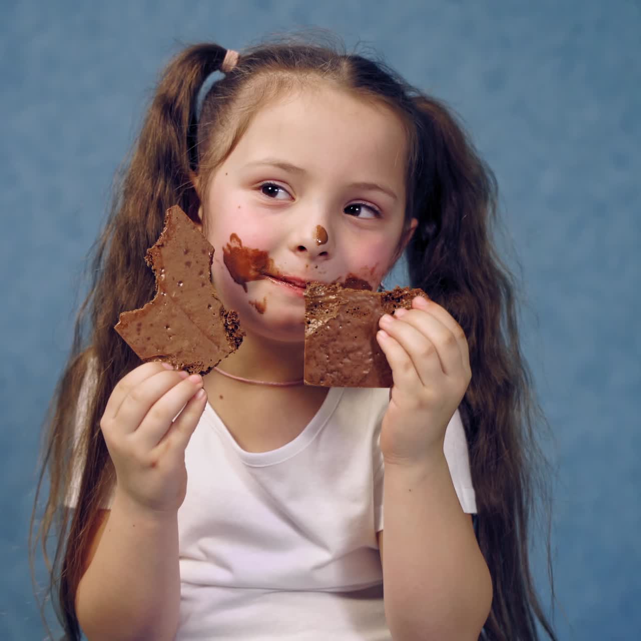 Portrait of a little girl eating chocolate. Funny kid with ponytails eats sweets with smeared face. Dirty child on blue studio background.