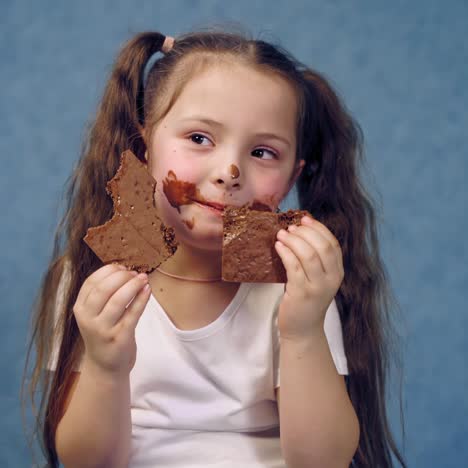 portrait of a little girl eating chocolate. funny kid with ponytails eats sweets with smeared face. dirty child on blue studio background.