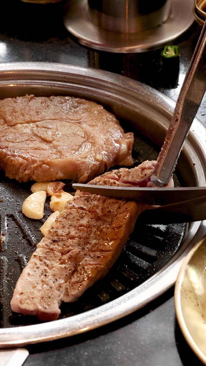 Pinze affettano una bistecca di maiale alla griglia su una griglia coreana da tavolo per barbecue con illuminazione calda e focalizzata
