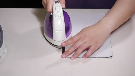 Hands-using-hot-iron-to-press-a-fold-on-the-edge-of-white-and-brown-fabric-on-white-colored-table