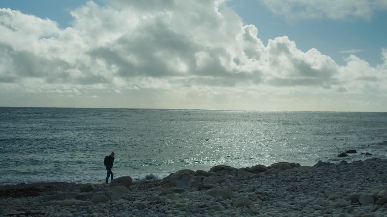 화창한 날 영국 도싯에 있는 포틀랜드 섬에 있는 교회 오페의 바위 해변을 걷고 있는 한 남자의 4k 슬로우 모션 파노라마 풍경 사진