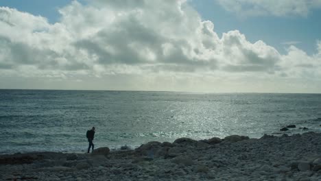 4K-Slow-motion-panoramic-landscape-shot-of-a-man-walking-on-the-rocky-beach-of-Church-Ope,-on-the-island-of-Portland,-in-Dorset,-England,-on-a-sunny-day