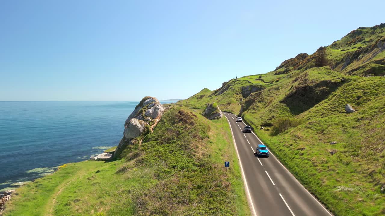 Aerial video of the Causeway Coastal Route in County Antrim, Northern Ireland, UK, on a bright and sunny day. Cars travel along the cliff-side road. Filmed in 4K, 60FPS and with Rec709 color.