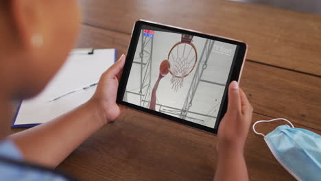 compuesto de una mujer sentada en una mesa con una máscara facial, viendo un juego de baloncesto en la tableta