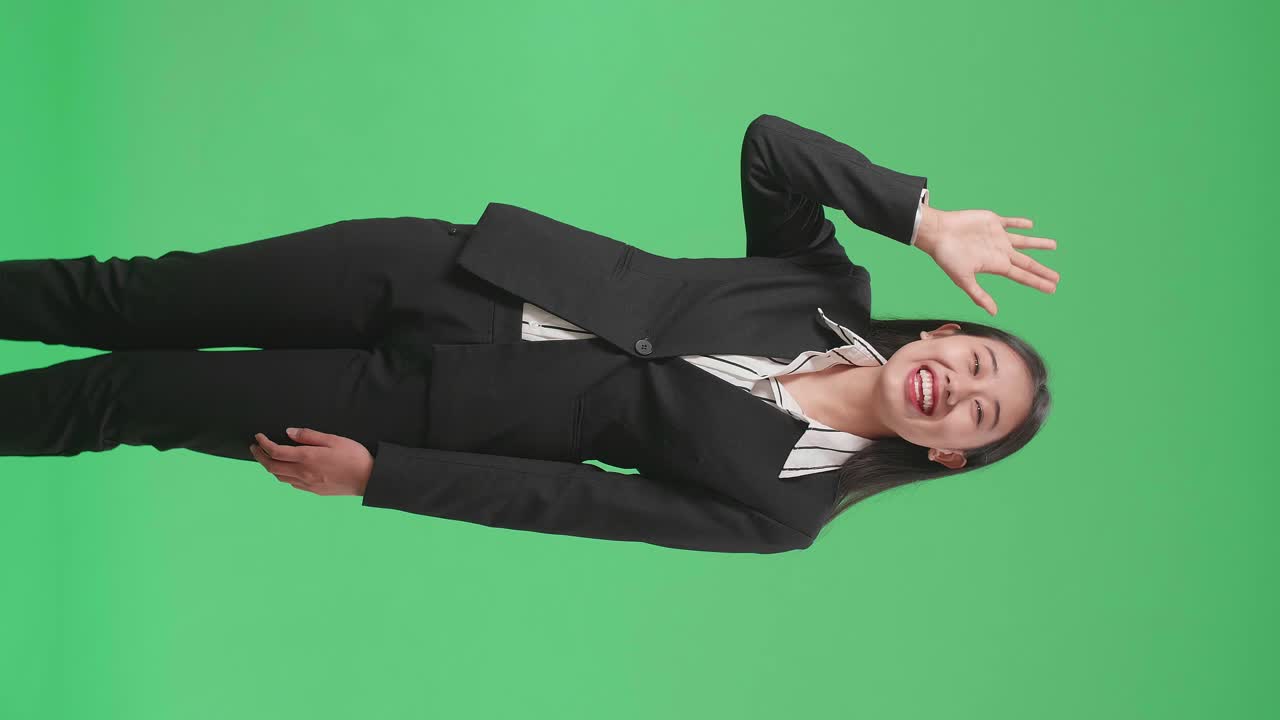 A Smiling Asian Business Woman Waving Hand And Say Hi While Standing On Green Screen In The Studio