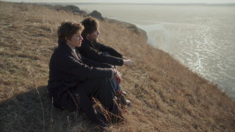 couple sitting on dry grassy slope and discussing scenic frozen lake