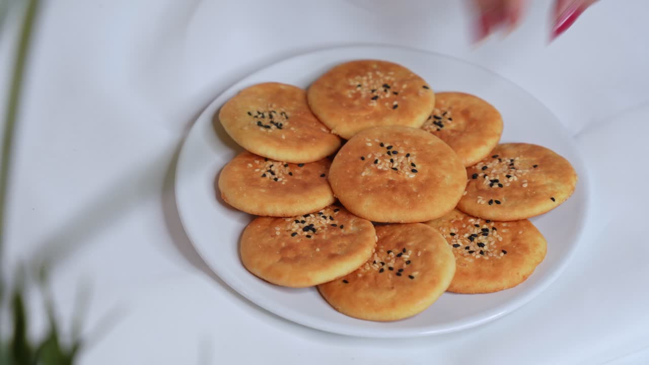 Close-up of a woman's hand picking a golden sesame cookie from a white plate. Traditional baked snack scene in 4K, ideal for food, culture, or lifestyle footage libraries.