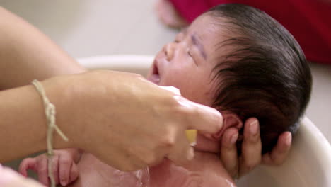 el cuello de un bebé que llora está siendo acurrucado mientras está a mitad de camino sumergido en una cuenca llena de agua, mientras sus padres le están dando un baño