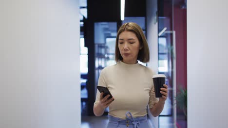 Mujer-De-Negocios-Asiática-Caminando-Usando-Un-Teléfono-Inteligente-Sosteniendo-Una-Taza-De-Café-En-Una-Oficina-Moderna