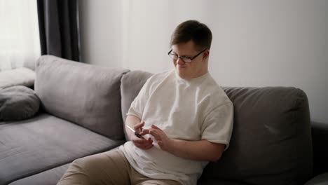 Smiling-Downs-Syndrome-guy-sitting-on-sofa-using-mobile-phone,-scrolling-internet