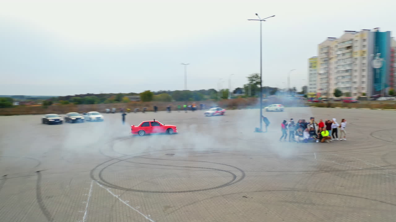 Red car riding circles drifting at the square. Bunch of people dancing hip hop at backdrop. High angle view.