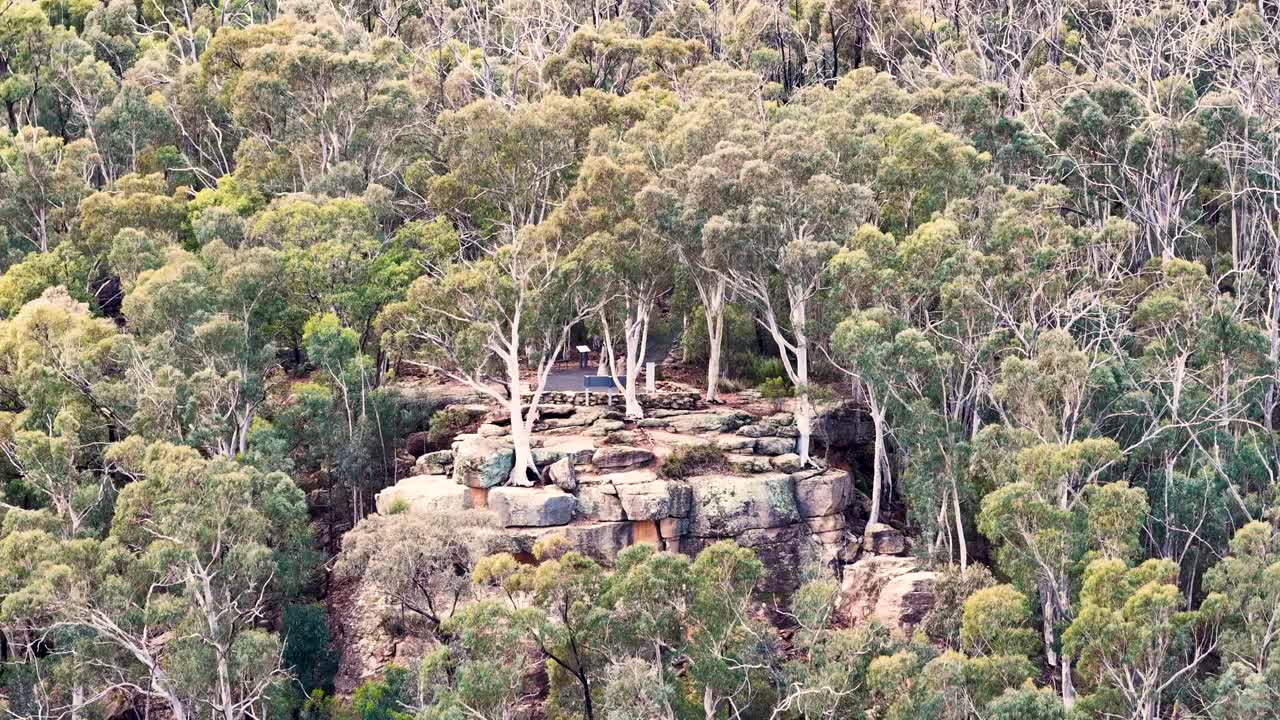 Aerial camera smoothly descends over a rocky sandstone outcrop surrounded by dense eucalyptus forest in soft daylight, revealing rugged terrain and native vegetation