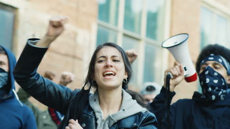 Close-up-view-of-caucasian-woman-in-facial-mask-yelling-with-arms-up-in-a-protest-with-multiethnic-group-of-people-in-the-street