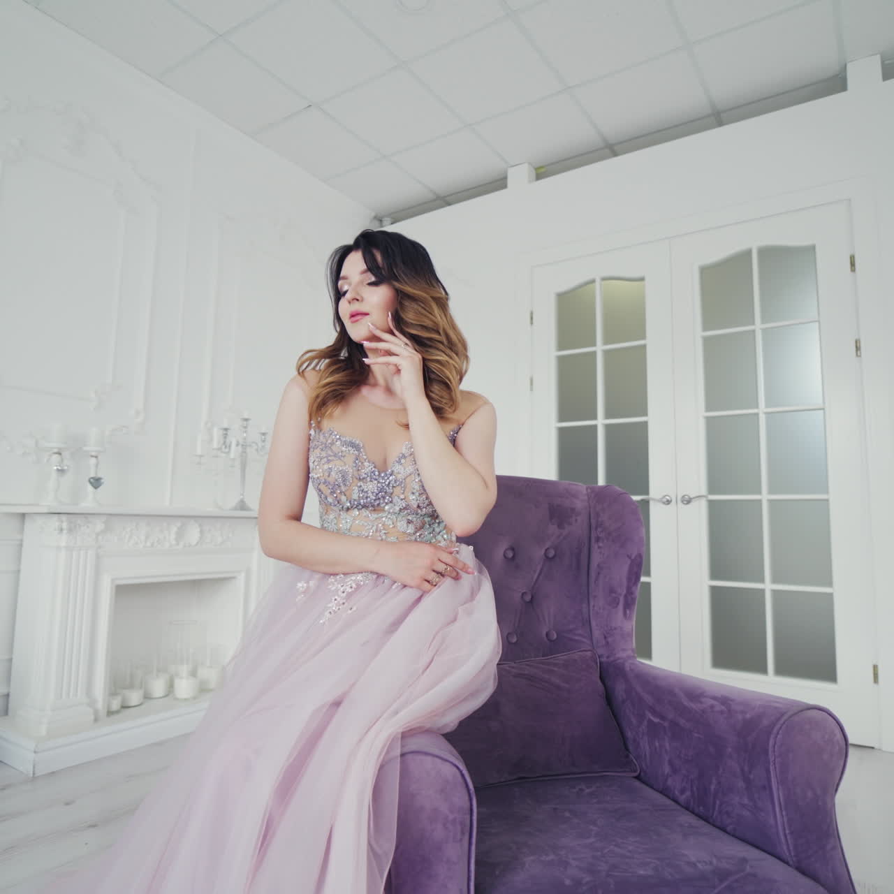 Beautiful bride in wedding dress admires herself while sitting on the chair in a room. Tendy model in a pretty dress is posing in a light studio on a violet armchair.