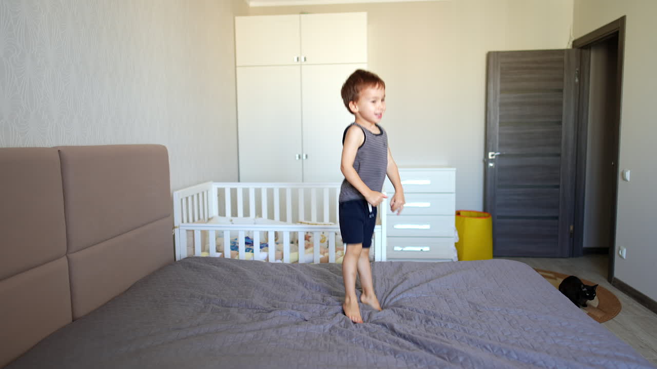 Cute Caucasian baby boy jumps on the big double bed. Little kid gets into his crib and hides there. Black cat sits on the floor.