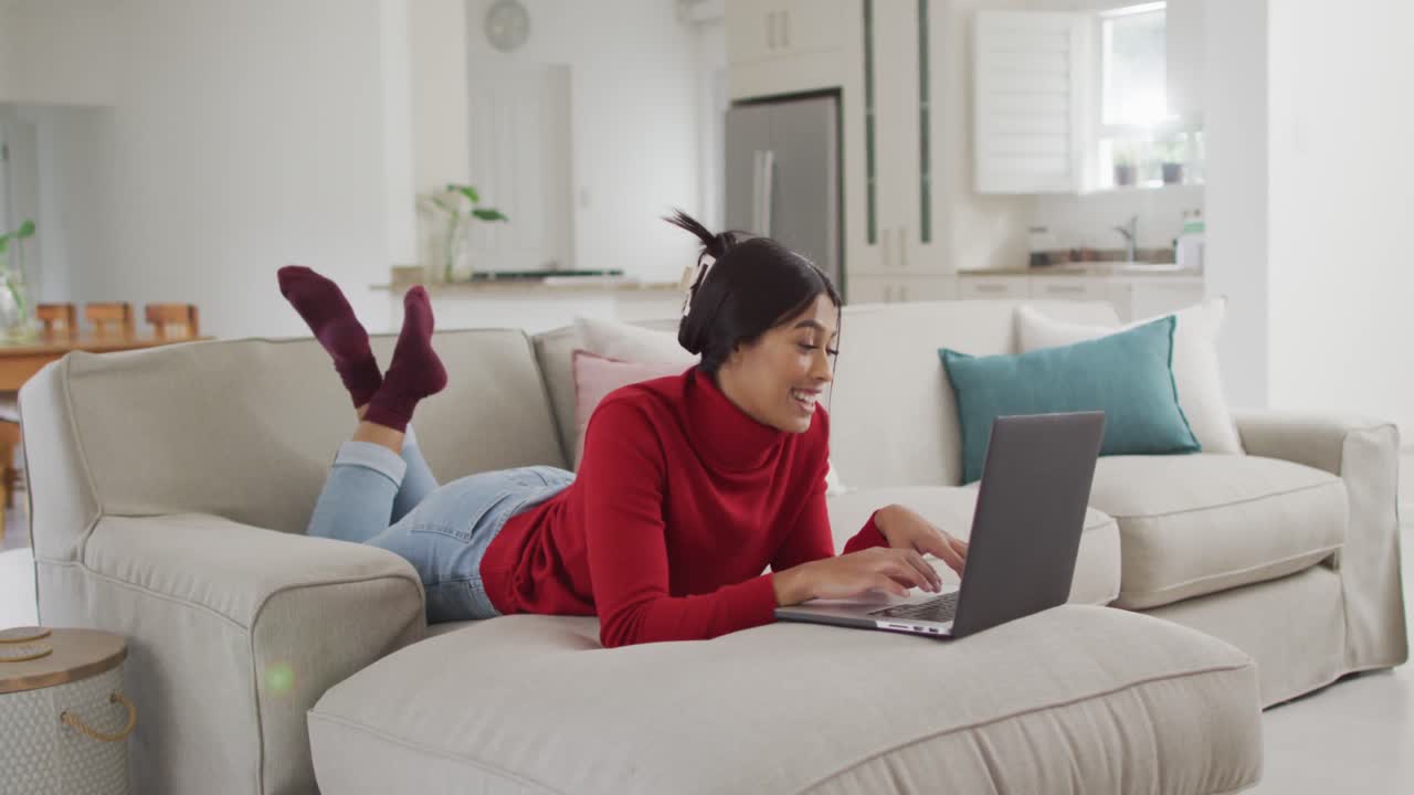 video de una feliz mujer biracial en hijab acostada en el sofá y usando una computadora portátil