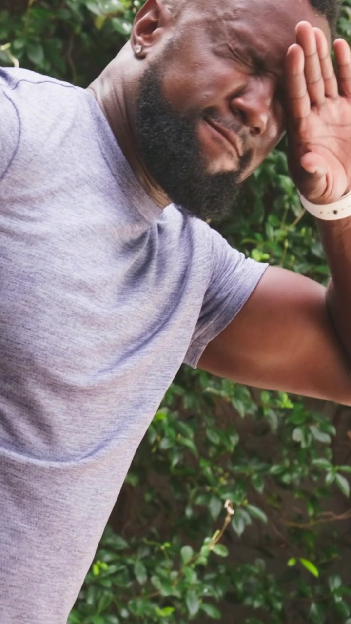 Vertical video: Sun hitting man wearing grey tee, shielding eyes and rubbing forehead by leafy wall
