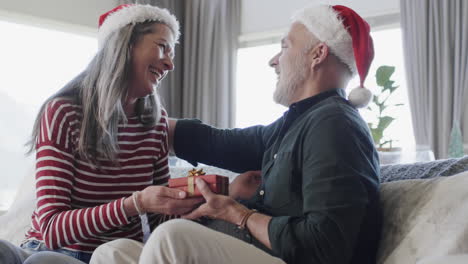 middle aged caucasian couple in santa hats gifting at christmas at home, slow motion