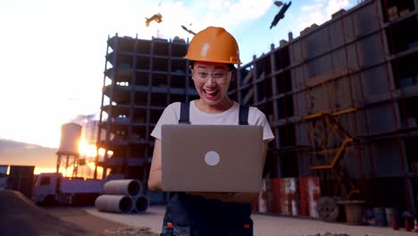 trabajadora asiática con gafas de protección y casco de seguridad mirando la computadora portátil y luego gritando meta celebrando mientras está de pie en el sitio de construcción