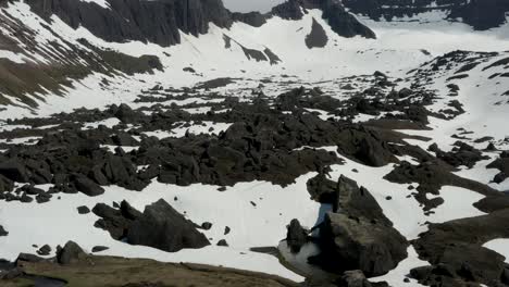 Amazing-tilt-up-from-Snow-covered-rocks-reveal-snow-capped-mountains-covered-in-clouds-on-a-sunny-day,-Iceland