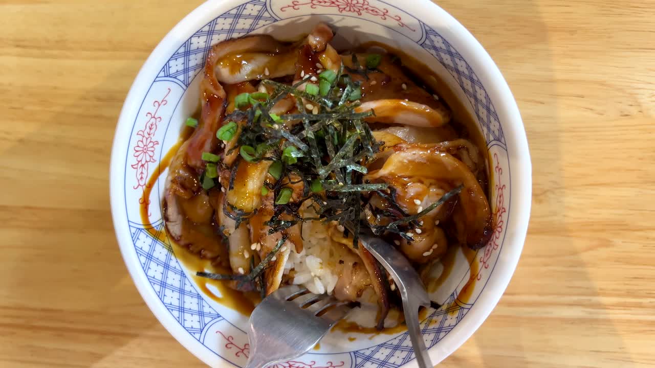 Hand uses spoon and fork to scoop grilled pork rice bowl under bright, natural lighting