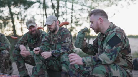 military men in camouflage uniforms resting after the afternoon shift at base in the field, sitting on crates outside and preparing a bonfire, putting sausages on sticks, drinking alcohol from a flask