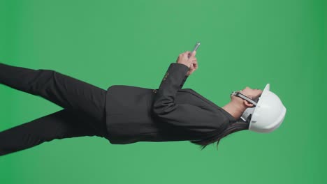 side view of smiling asian female engineer looking around and shaking her head while using smartphone and walking in the green screen studio