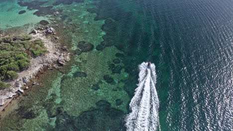 A-speedboat-cruising-over-clear-turquoise-waters-near-a-rocky-coastline-in-sardinia,-aerial-view