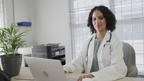 portrait of biracial female doctor wearing lab coat, sitting at desk and using laptop, slow motion