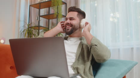 feliz y relajado joven caucásico con auriculares inalámbricos escuchando música usando una computadora portátil en el sofá