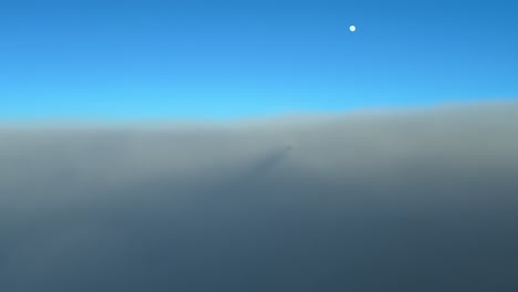 aerial view taken from inside a jet cockpit and its chemtrail while flying at sunset over a sea of ethereal clouds, with the moon above in a blue sky