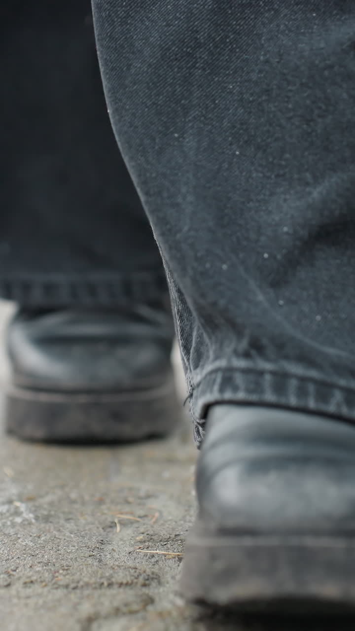Primo piano dei pantaloni neri e degli stivali neri di qualcuno che cammina lentamente in avanti su un sentiero pavimentato all'aperto durante una giornata calma e nuvolosa, concentrandosi sul movimento delle calzature, il ritmo dei passi e l'atmosfera fresca.